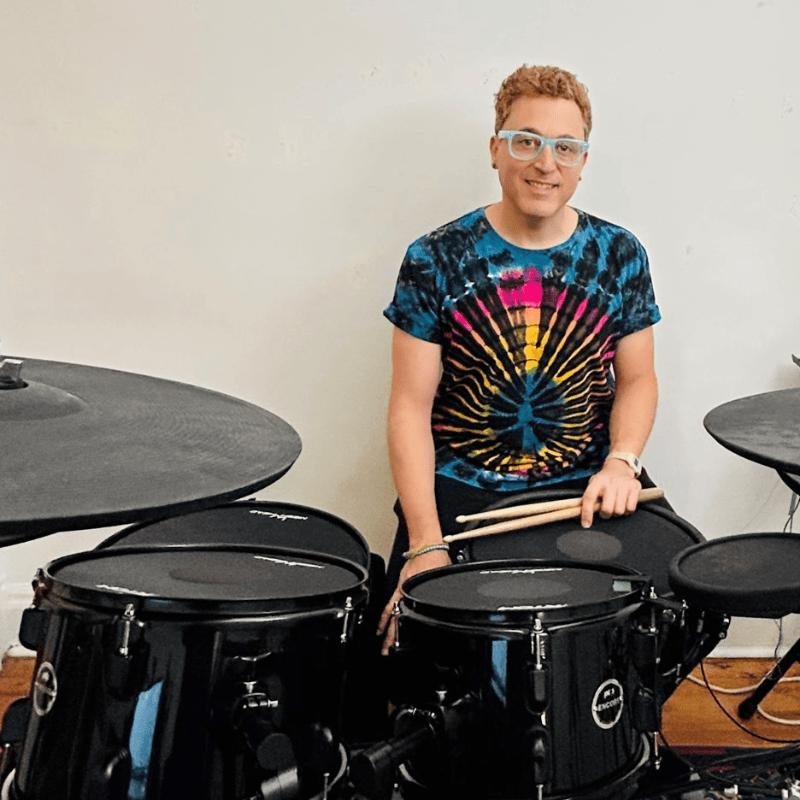 Person wearing a tie-dye shirt with drum set in front of a white wall