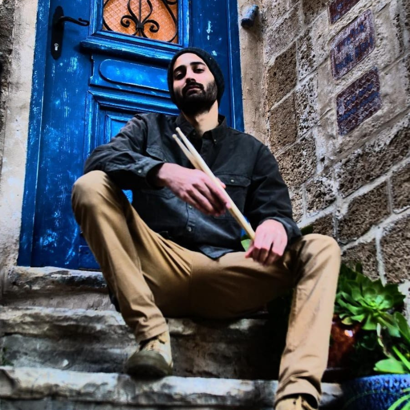 Man sitting on stone steps holding drumsticks in front of a blue door.